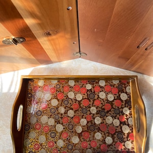 May include: A rectangular wooden serving tray with a floral pattern. The tray has a dark brown border and handles. The floral pattern features red, white, and gold flowers on a dark brown background. The tray is likely used for serving food or drinks.