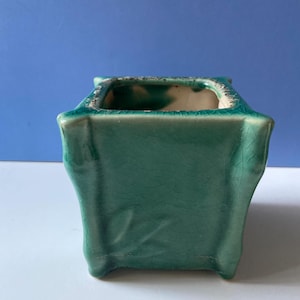 May include: A square, teal-colored ceramic bonsai pot with a textured, crackled glaze. The pot has a slightly flared shape and a white interior. The corners are slightly rounded, and the pot sits on four small feet.