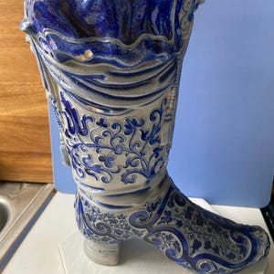 Może przedstawiać: Ozdobny ceramiczny wazon w kształcie buta w odcieniach niebieskiego i szarego. But ma skomplikowane wzory kwiatowe i zwoje. Górna krawędź ma falisty wzór. Wazon to unikalny element wystroju domu.
