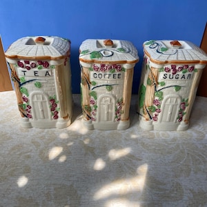 Pode incluir: Conjunto de três recipientes de cerâmica para chá, café e açúcar. Cada recipiente tem a forma de uma pequena casa com uma porta branca, um telhado e detalhes florais. As palavras "TEA", "COFFEE" e "SUGAR" estão impressas na frente.