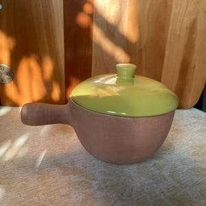 May include: A terracotta casserole dish with a green lid and a handle. The dish is on a wooden surface.
