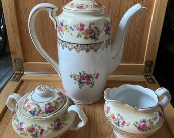 May include: A porcelain tea set consisting of a tall teapot with a lid, a lidded sugar bowl, and a creamer. Each piece features a cream-coloured base, a floral pattern of pink and yellow roses with blue accents, and gold trim detailing.