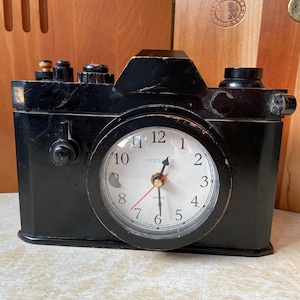 Puede incluir: Un reloj de cuarzo negro con forma de cámara, esfera blanca, números negros y segundero rojo. El reloj presenta una esfera circular similar a un objetivo, botones en la parte superior y el texto "MODERNIZE" y "QUARTZ" impreso en el centro.