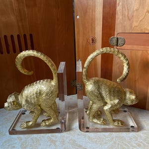 May include: A pair of gold-colored monkey bookends. Each monkey is in a crouching position with a curled tail. The bookends are mounted on clear acrylic bases, adding a touch of elegance to any bookshelf or desk.