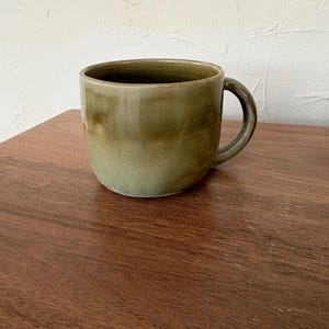 May include: A ceramic mug with a rounded body and a handle. The mug has a gradient of olive green hues, with a darker shade at the rim and a lighter shade towards the base. It sits on a wooden surface.