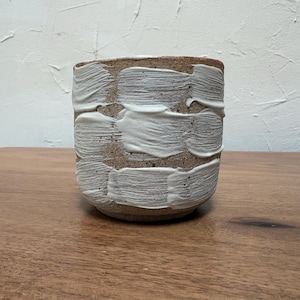 May include: A cylindrical ceramic planter featuring a speckled tan clay base with thick, horizontal white paint strokes applied in a textured, layered pattern around the exterior surface, creating a tactile and dimensional appearance.