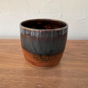 May include: A handmade ceramic bowl with a glossy finish. The bowl features a gradient of colors, transitioning from a dark brown top to a teal-blue middle and a reddish-brown base. The bowl is sitting on a wooden surface.