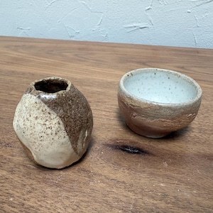 May include: A small, round, textured ceramic vase with a speckled cream and brown finish sits beside a wide, shallow ceramic bowl featuring a speckled white interior and a natural, earthy-toned exterior with a rough, hand-formed clay texture.
