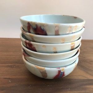 May include: A stack of six white ceramic bowls with a light blue glaze and brown and red abstract patterns.