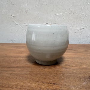 May include: A rounded ceramic bowl with a glossy off-white glaze and subtle speckled texture. The vessel features a wide, open mouth and a slightly tapered base, showcasing a smooth, curved silhouette with faint horizontal indentations.