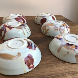 May include: Six ceramic bowls with a white glaze and brown, red, and gold abstract patterns. The bowls are arranged in a row on a wooden surface.