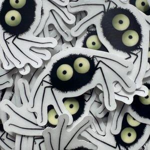 May include: A collection of white stickers featuring a cartoonish creature with a black, fuzzy body and large, yellow eyes. The creature has four white, hand-like appendages. The stickers are scattered, creating a textured, layered effect.