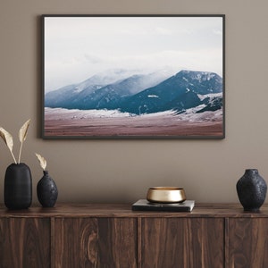 May include: A framed photograph of a mountain range with snow-capped peaks and a misty sky. The mountains are a deep blue color, and the sky is a light blue. The foreground is a field of brown grass.