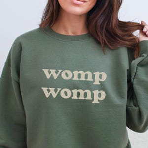 May include: Olive green sweatshirt with the text "womp womp" in a light beige color. The sweatshirt has a crew neck and long sleeves. The image is a close-up of the upper body.