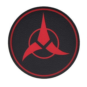 May include: A round black patch with a red emblem. The emblem features a stylized three-pointed shape within a red circle, and a red outline. The patch has a textured appearance and is set against a white background.