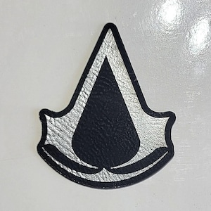 May include: A black and silver emblem with a stylized design. The emblem features a black teardrop shape within a silver border, with a textured appearance. The emblem is likely a logo or patch.