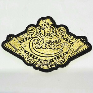 May include: A gold and black decorative patch with the text "Auntie's Choice Co." The patch features a woman's face and ornate scrollwork. The patch has a glittery gold finish and a black border.