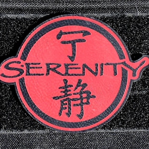 May include: A circular patch featuring a red background with a black border. The word SERENITY is printed in black across the center. Above the text is a black Chinese character, and below the text is another black Chinese character.