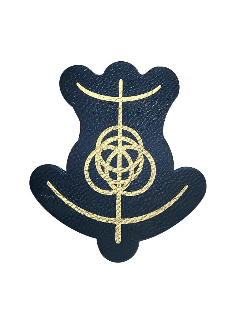 Op de afbeelding: Een marineblauwe leren patch met een goudkleurig geometrisch ontwerp. Het ontwerp heeft een centrale verticale lijn die wordt gekruist door horizontale lijnen en onderling verbonden cirkels. De patch heeft een unieke, gestileerde vorm.