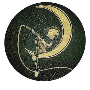 Pode incluir: Base redonda verde-escura com uma lua crescente dourada e uma figura fantasiosa. A figura está sentada na lua, segurando uma vara de pescar com uma linha de folhas. A base tem uma superfície texturizada.