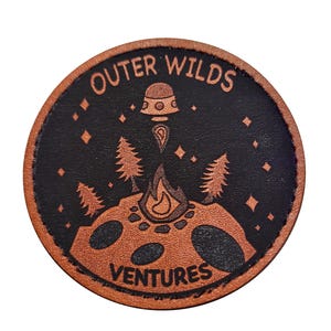 May include: A circular, brown and black leather patch with the words "Outer Wilds Ventures" in a circular design. The patch features a stylized illustration of a campfire, trees, and a spaceship. The background is black with small diamond shapes.