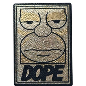Könnte beinhalten: Ein rechteckiger Aufnäher mit einem Gold- und Schwarz-Design. Der Aufnäher zeigt ein Cartoon-Gesicht mit grimmigem Ausdruck und dem Wort "DOPE" in fetten schwarzen Buchstaben. Das Gesicht hat ein strukturiertes Aussehen.