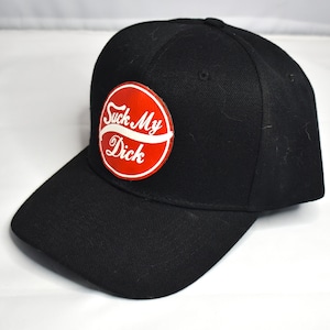 May include: Black baseball cap with a red circular patch. The patch features the text "Suck My Dick" in white script. The cap has a curved brim and a structured crown.