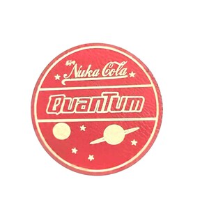 May include: A round, red Nuka Cola Quantum design with gold lettering and accents. The design includes the text "Nuka Cola Quantum" and a stylized planet and stars.