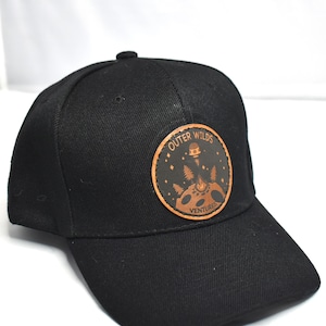 May include: Black baseball cap with a curved brim. The cap features a circular patch with the words "Outer Wilds Ventures" and a graphic of a campfire, trees, and a planet. The patch is brown and the cap is black.
