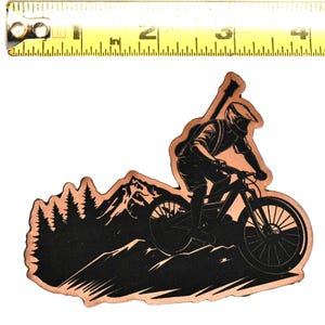 May include: A copper-colored sticker featuring a black silhouette of a mountain biker riding on a mountain bike. The sticker is approximately 3 inches long, with a mountain range and trees in the background.