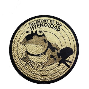 Puede incluir: Un parche redondo de color dorado con un borde negro que presenta una ilustración estilizada de una rana con collar. El texto "ALL GLORY TO THE HYPNOTOAD" está impreso alrededor del borde del parche.