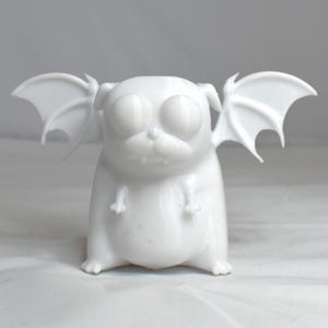 Peut inclure: Figurine blanche représentant un carlin avec de grands yeux et des ailes de chauve-souris. Le carlin est assis, ses pattes avant sont visibles. Les ailes sont déployées sur les côtés. La figurine est sur une surface blanche.