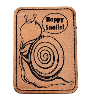 Puede incluir: Un parche de cuero marrón con una ilustración negra de un caracol de dibujos animados y el texto "Happy Snails!". El caracol tiene una concha grande y está sonriendo. El parche tiene un borde cosido.