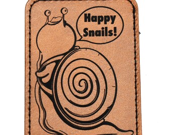 May include: A tan leather patch with a black illustration of a cartoon snail and the text "Happy Snails!". The snail has a large shell and is smiling. The patch has a stitched border.