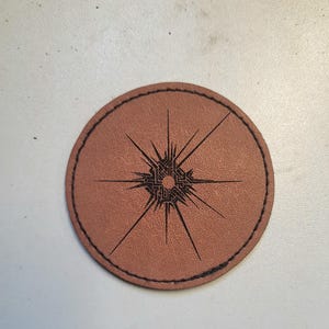 Może przedstawiać: Brązowy podkładka pod kubek ze skóry z czarnym wzorem w kształcie gwiazdy. Wzór jest skomplikowany i szczegółowy, z centralnym okręgiem i promieniującymi liniami.