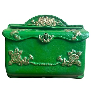 May include: A green decorative box with gold floral detailing. The box has a curved top with a floral design, a central clasp, and floral embellishments on the front and sides. The box is painted in a glossy green.