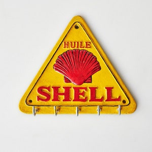May include: Yellow triangular key holder with the Shell logo in red. The key holder has the word "SHELL" in red block letters and the word "HUILE" above the logo. It features five silver hooks for keys.