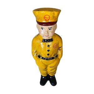 May include: A yellow ceramic figurine of a Shell gas station attendant. The figurine is wearing a yellow uniform with black buttons, a black belt, and a black cap with a red Shell logo. The figurine is standing with its hands in its pockets.