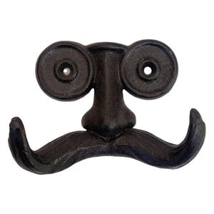 May include: A dark brown cast iron wall hook shaped like a face with a prominent nose, two circular eye-like mounting holes, and a wide, curved mustache that serves as the double hook for hanging items. The metal has a textured, matte finish.