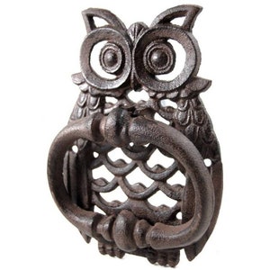 May include: A cast iron owl-shaped door knocker in a rustic brown finish. The knocker features a large ring for knocking and intricate details, including the owl's eyes, feathers, and a decorative pattern below the ring. The door knocker measures approximately 15 cm in height.