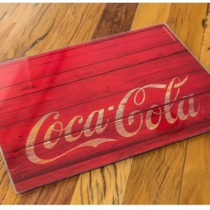 May include: Red wooden cutting board with the Coca-Cola logo in gold lettering.