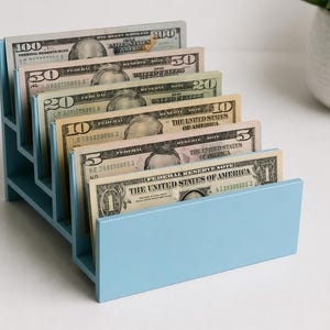 May include: A light blue tiered wooden organizer holding United States paper currency in descending denominations from one hundred dollars to one dollar, arranged in separate slots to display each bill clearly in a stepped, cascading formation.