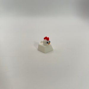 May include: White keycap with a small white chicken figurine on top. The chicken has red wattles and a yellow beak.