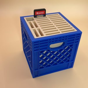 May include: A blue and white 3D-printed crate with a Nintendo Switch game cartridge on top. The crate has a grid-like top and a rectangular shape with cut-out handles on the sides. The game cartridge is black and red.