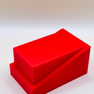 May include: A bright red, geometric sculpture composed of two stacked rectangular blocks. The top block is angled, creating a dynamic visual effect. The piece is set against a plain white background, highlighting its form and color.
