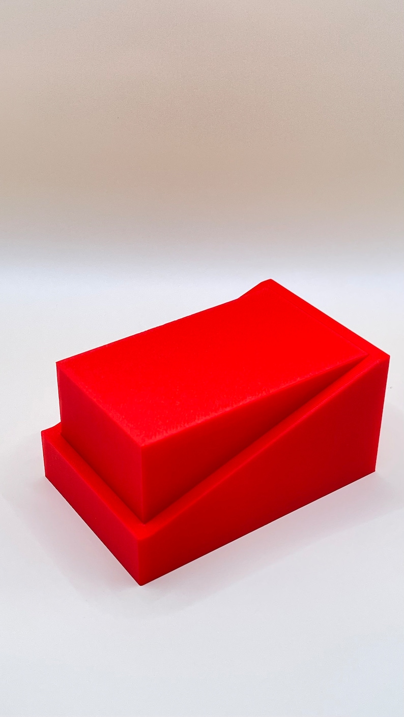 May include: A bright red, geometric sculpture composed of two stacked rectangular blocks. The top block is angled, creating a dynamic visual effect. The piece is set against a plain white background, highlighting its form and color.