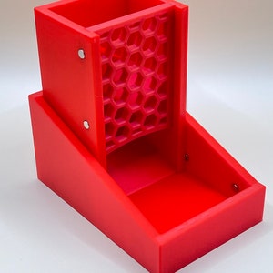 May include: A vibrant red dice tower featuring a honeycomb-patterned dice chute. The tower is rectangular with a tray at the base. The tower is made of plastic and has a modern design.