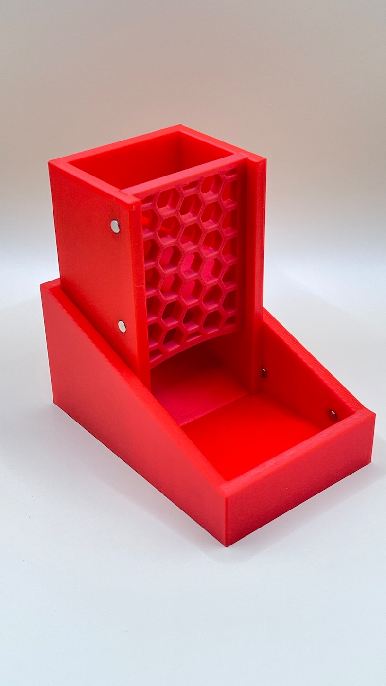 May include: A bright red dice tower with a honeycomb-patterned dice chute. The tower is rectangular with a tray at the base. The tower is made of plastic and has a modern design.