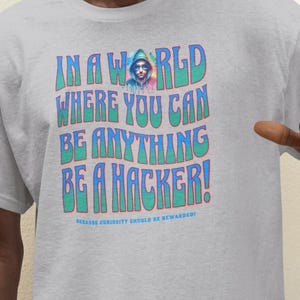 Könnte beinhalten: Ein graues T-Shirt mit einer bunten Grafik, auf der "In einer Welt, in der du alles sein kannst, sei ein Hacker!" steht. Die Grafik zeigt eine Person mit Kapuzenpullover und Sonnenbrille.