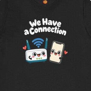 May include: A black short-sleeve t-shirt featuring a graphic of a smiling Wi-Fi router and a smartphone with the text We Have a Connection. The design includes blue Wi-Fi signal waves and small red hearts surrounding the two cartoon characters.